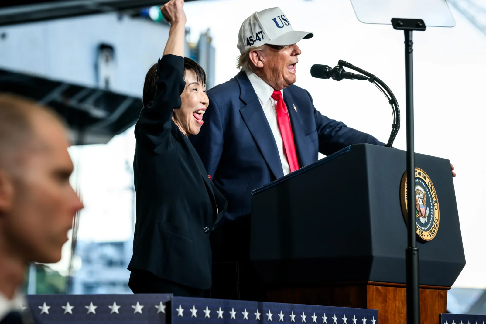 President Trump and Japanese Prime Minister Sanae Takaichi stand at a podium together, as Takaichi waves.