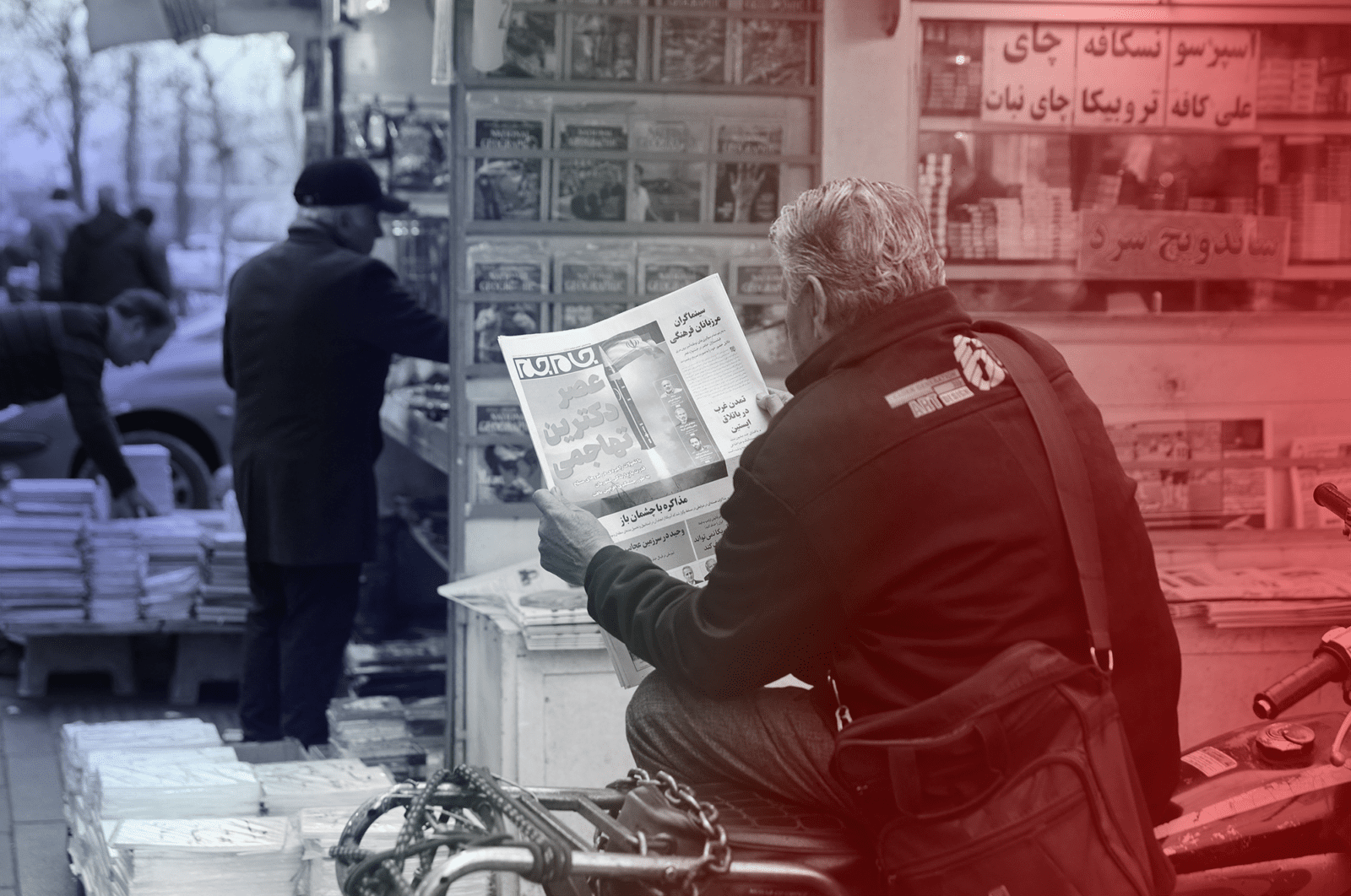 A man sits beside a newsstand reading a paper in Farsi which shows a missile.