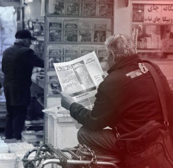 A man sits beside a newsstand reading a paper in Farsi which shows a missile.