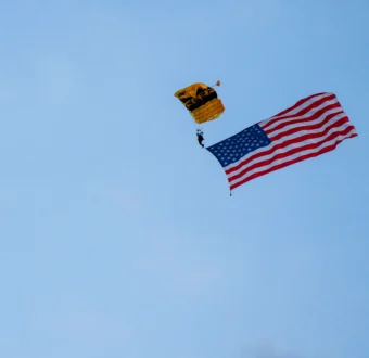 An Army paratrooper descends through the sky with a flying American flag.