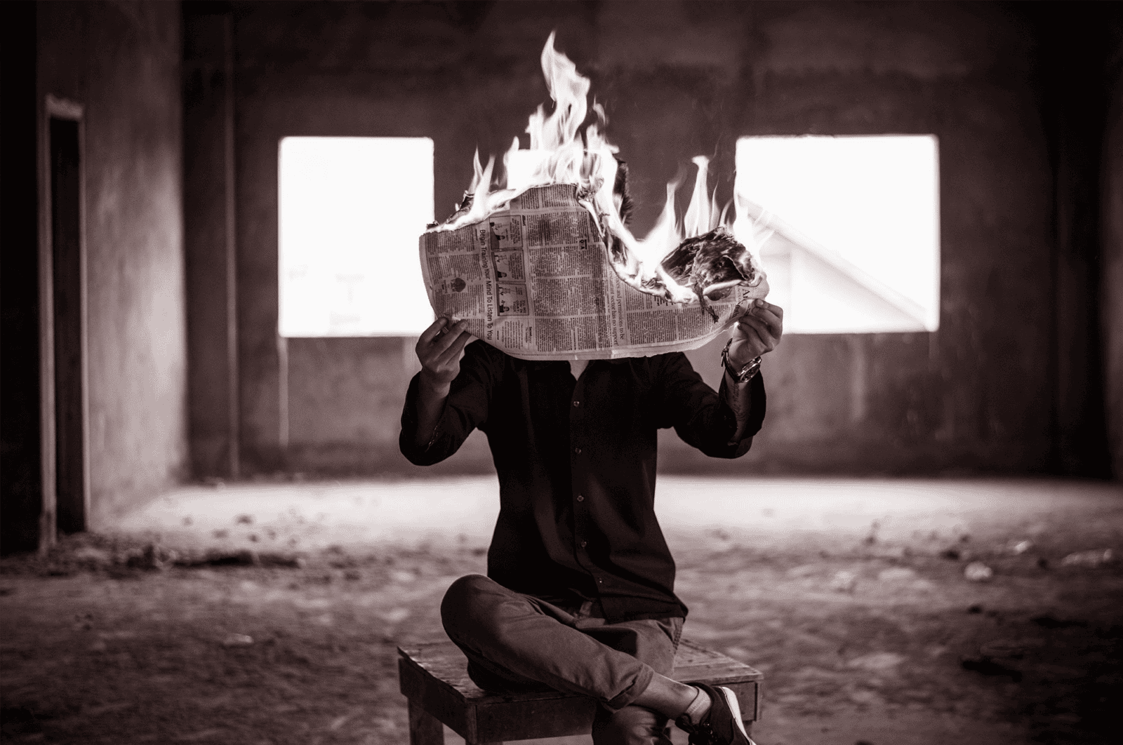 A man in an abandoned building sits holding a burning newspaper.