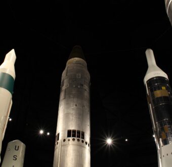 Looking up at a ring of tall ballistic missiles under a dark hangar ceiling dotted with lights, one marked "USA."