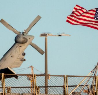 The tail rotor of a gray Navy helicopter rises behind a deck fence beside an American flag streaming from a flagpole against a clear blue sky.
