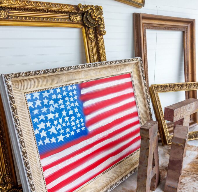 A hand-painted American flag reading "God Bless America" in an ornate frame, propped among empty gilded frames in a gallery window.