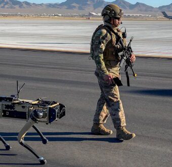 A US service member in a combat helmet, carrying a rifle, walks across an airfield apron beside a four-legged robot with a radio antenna, with desert mountains and a transport plane behind them.
